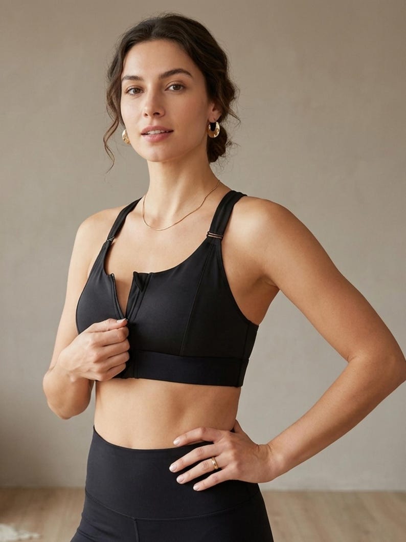 Op de afbeelding: Een zwarte sport-bh met een ritssluiting aan de voorkant. De bh heeft verstelbare bandjes en een ondersteunend ontwerp. Het model draagt de bh met zwarte leggings. De achtergrond is een neutrale kleur.