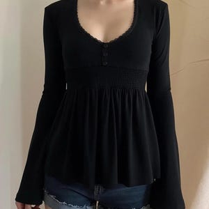 May include: Black long-sleeved top with a scoop neckline trimmed with lace. The top has three small buttons and a gathered waist. The sleeves are flared at the wrists. The top is paired with denim shorts.
