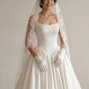 Op de afbeelding: Een witte trouwjurk met een vierkante halslijn, voorzien van gedetailleerd bloemenborduursel op het lijfje en een volle satijnen rok. De set bevat een lange, doorschijnende sluier met kanten rand, witte satijnen handschoenen, een parelketting en pareloorbellen.