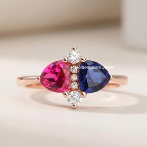 Puede incluir: Un anillo de oro rosa con un diseño llamativo. Presenta una gema rosa fucsia y una gema azul oscuro, ambas triangulares. Pequeñas gemas redondas y claras están engastadas entre las piedras más grandes. El anillo está sobre un fondo suave y neutro.