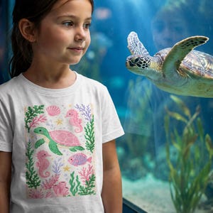 Könnte beinhalten: Ein weißes Kurzarm-T-Shirt mit einem farbenfrohen Grafikdruck einer grünen und rosafarbenen Meeresschildkröte, zwei rosafarbenen Seepferdchen, einem kleinen rosa Fisch, grünem Seetang, rosa Korallen und gelben Seesternen.