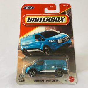 May include: A Matchbox die-cast toy of a 2023 Ford E-Transit Custom van. The van is light blue with black wheels and white accents. The packaging is red and features the Ford logo and the Matchbox logo.