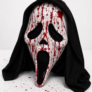 May include: A white plastic ghost mask featuring a long, distorted face with large black eye sockets and an elongated open mouth. The mask is covered in red paint splatters resembling blood and is attached to a black fabric hood that drapes down.