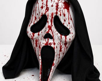 May include: A white plastic ghost mask featuring a long, distorted face with large black eye sockets and an elongated open mouth. The mask is covered in red paint splatters resembling blood and is attached to a black fabric hood that drapes down.
