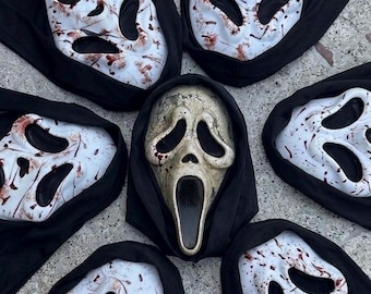May include: A collection of six white plastic masks with black hoods, featuring elongated eye and mouth openings. Five masks are splattered with red paint, while the central mask has a weathered, aged, and textured off-white finish.