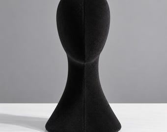 May include: A black velvet-covered mannequin head display stand with a smooth, featureless oval face and a flared base. The surface has a soft, matte texture, and a vertical seam runs down the centre of the face and neck area.