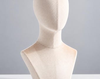 May include: A beige velvet mannequin head and shoulder bust with a smooth, featureless face and a vertical seam running down the centre of the head. The display form has a rounded neck and a wide base, designed for showcasing hats or accessories.