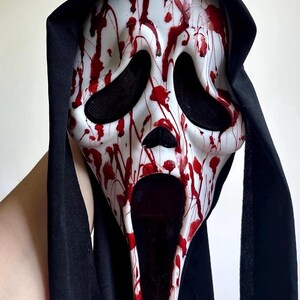 May include: A white plastic mask featuring a distorted, elongated face with large black eye sockets and an open mouth. The mask is covered in red paint splatters resembling blood and is attached to a black fabric hood that drapes down the sides.