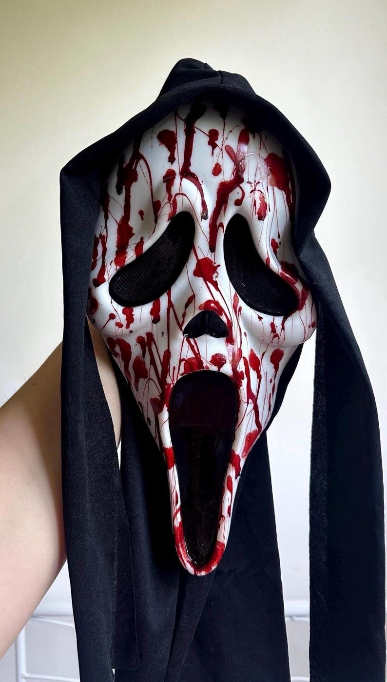 May include: A white plastic mask featuring a distorted, elongated face with large black eye sockets and an open mouth. The mask is covered in red paint splatters resembling blood and is attached to a black fabric hood that drapes down the sides.