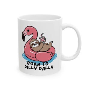 May include: White ceramic mug featuring a cartoon sloth relaxing on a pink flamingo float with a drink. The mug has the text "BORN TO DILLY DALLY" in black letters. The design includes blue water details.