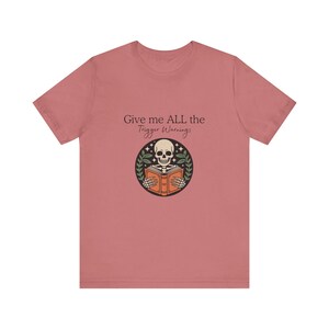 Può includere: Una t-shirt a maniche corte color rosa polvere con una grafica di uno scheletro che legge un libro arancione all'interno di un cerchio nero. Sopra la grafica, il testo recita Give me ALL the Trigger Warnings in un carattere marrone scuro, serif e corsivo.