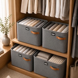 Peut inclure: Quatre bacs de rangement rectangulaires en tissu gris avec des coutures blanches et des poignées en similicuir beige. Chaque bac comporte une petite étiquette noire avec le mot Fashion imprimé en blanc. Les bacs contiennent des textiles pliés.