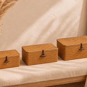 May include: Three rectangular woven storage boxes in graduated sizes. The boxes are a warm tan colour, with a textured surface and a dark wooden clasp. These boxes are suitable for home decor and are made of natural materials.
