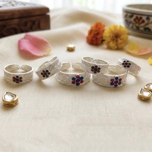 May include: Silver adjustable rings with floral designs and colorful gemstones. The rings are arranged on a cream-colored surface, surrounded by flower petals and small gold-rimmed gems. A wooden box and a decorative bowl are in the background.