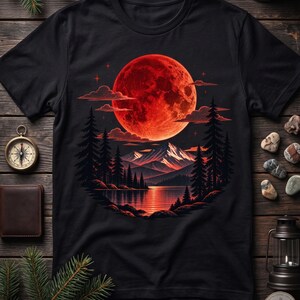 Könnte beinhalten: Ein schwarzes Kurzarm-T-Shirt mit einem kreisförmigen Grafikdruck, der einen großen roten Mond über einem dunklen Gebirge, einem ruhigen See und einem Kiefernwald zeigt. Das Design verwendet Rot-, Orange- und Schwarztöne für eine nächtliche Szene.