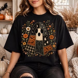 Puede incluir: Una camiseta negra de manga corta con un gran gráfico central de un fantasma de patchwork. El diseño del fantasma incorpora varios patrones, incluyendo cuadros negros y blancos, estampados de murciélagos naranjas y motivos florales oscuros, rodeado de flores.