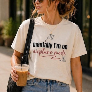 May include: Cream-colored t-shirt with the text "mentally I'm on airplane mode" in black and burgundy. The shirt features a small airplane graphic and the words "NO SIGNAL". The person is wearing blue jeans and sunglasses.