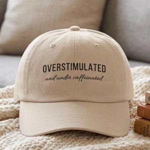 May include: A beige baseball cap with the words "OVERSTIMULATED and under caffeinated" in black script. The hat is sitting on a cream-colored blanket, with a couch and books in the background.