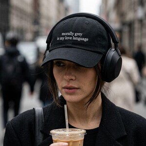 May include: Black baseball cap with the text "morally grey is my love language" in white. The cap is worn by a person wearing black headphones and a black coat. The person is holding a clear plastic cup with a straw.