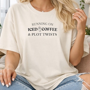 Può includere: T-shirt color crema con la scritta nera "RUNNING ON ICED COFFEE & PLOT TWISTS". Il design include una grafica di caffè freddo. La maglietta è in stile casual e oversize.