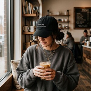May include: Black baseball cap with the text "morally grey is my love language" in white. The person is wearing a gray sweatshirt and holding a glass of iced coffee. The background shows a coffee shop setting with bookshelves and other people.