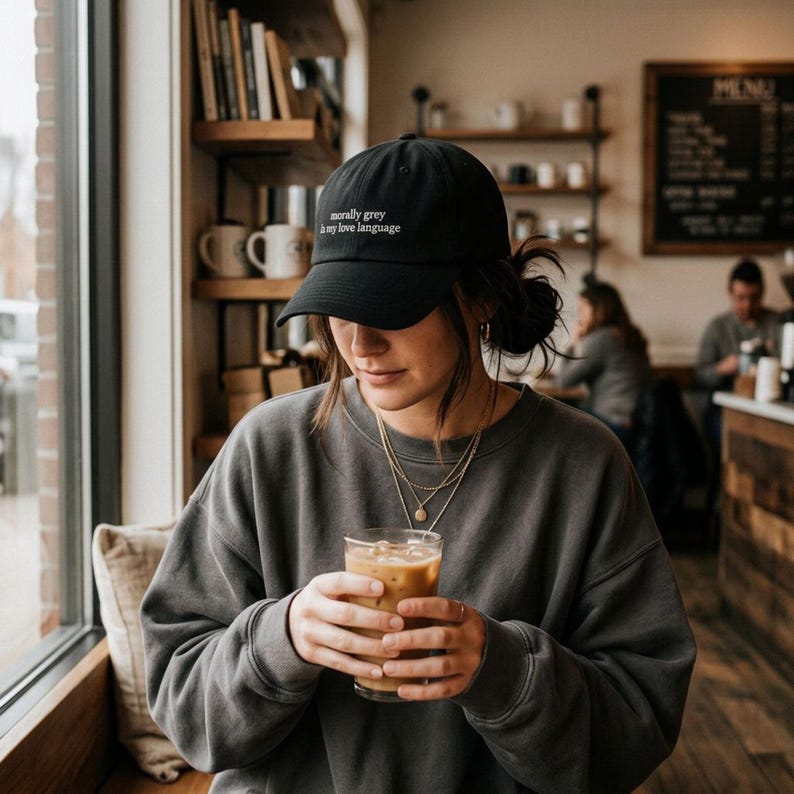 May include: Black baseball cap with the text "morally grey is my love language" in white. The person is wearing a gray sweatshirt and holding a glass of iced coffee. The background shows a coffee shop setting with bookshelves and other people.