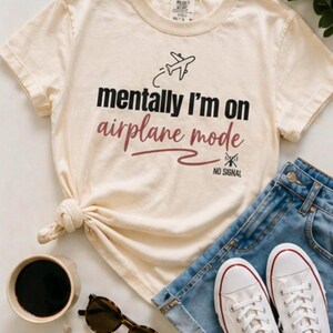 May include: A cream-coloured t-shirt with the text "mentally I'm on airplane mode" and an airplane graphic. The shirt is styled with denim shorts, white trainers, sunglasses, and a coffee cup.