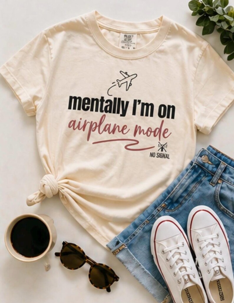 May include: A cream-coloured t-shirt with the text "mentally I'm on airplane mode" and an airplane graphic. The shirt is styled with denim shorts, white trainers, sunglasses, and a coffee cup.