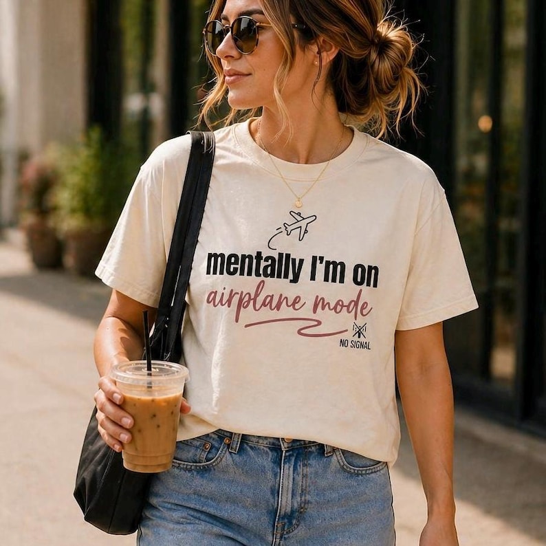 May include: Cream-colored t-shirt with the text "mentally I'm on airplane mode" in black and burgundy. The shirt features a small airplane graphic and the words "NO SIGNAL". The person is wearing blue jeans and sunglasses.