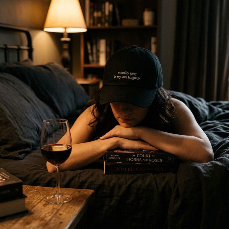 May include: A black baseball cap with the text "morally grey is my love language" in white. A glass of red wine sits on a wooden table next to a stack of books. The scene is set in a bedroom with a lamp.