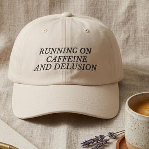 May include: A beige baseball cap with the text "RUNNING ON CAFFEINE AND DELUSION" in black. The cap is on a neutral-colored surface, with a coffee cup and lavender sprigs in the background.