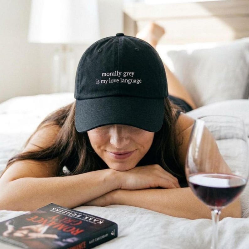 May include: Black baseball cap with the text "morally grey is my love language" in white. The cap is worn by a person lying on a bed. A book and a glass of red wine are also visible.
