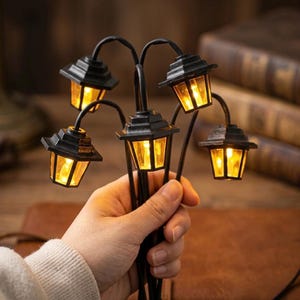 Op de afbeelding: Een cluster van miniatuur zwarte straatlantaarns met gloeiende gele lichten. Elke lamp heeft een vierkante, gelaagde bovenkant en een gebogen arm. De lampen worden in een hand gehouden, wat suggereert dat het een decoratief of ambachtelijk item is.