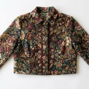 May include: A cropped jacket featuring a dark burgundy base with an intricate floral tapestry pattern in shades of green, cream, and muted red. The jacket includes a standard collar, long sleeves with buttoned cuffs, and a front button closure.