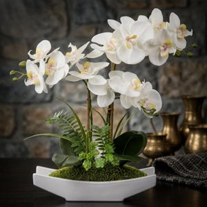 May include: A potted artificial orchid plant featuring multiple white flowers with pale yellow and pink centers. The plant includes green fern-like foliage and moss at the base, all arranged in a white, elongated, boat-shaped ceramic planter.