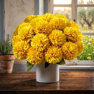 Puede incluir: Un denso ramo de flores de crisantemo de color amarillo vibrante dispuestas en un sencillo jarrón cilíndrico de cerámica blanca. Las flores presentan numerosos pétalos apretados, creando un aspecto redondeado y lleno con hojas verdes visibles en la base.