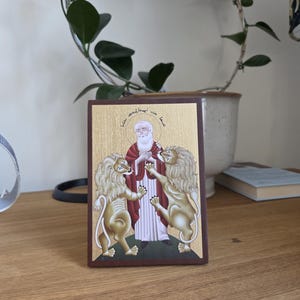 May include: A rectangular religious icon featuring a saint with a white beard and red robes, flanked by two golden lions. The background is gold with text at the top. The icon is set in a dark brown frame.