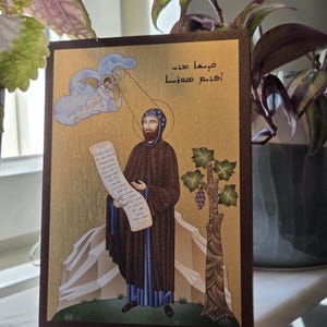 May include: A framed religious icon featuring a figure in brown robes holding a scroll, with a halo and a cloud with an angel. The background is gold with a tree and grapes. The frame is dark brown.