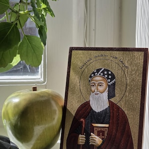 May include: A religious icon featuring a man with a white beard, wearing a black hat and red robe, holding a book and cane. The icon has gold accents and is set against a wooden frame. A green apple and plant are in the background.