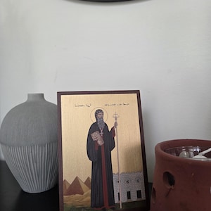 May include: A framed religious icon featuring a figure in a black robe holding a book and staff. The icon has a gold background with images of pyramids and a building. The frame is dark brown.