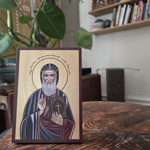 May include: A religious icon featuring a saintly figure with a long white beard, holding a book with a cross. The icon has a gold background and is framed in dark brown. The saint is wearing a black and red robe.