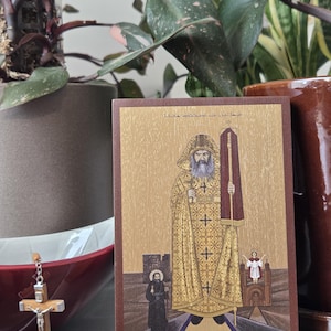 May include: A religious icon featuring a figure in a gold robe with a long white beard, holding a staff and a red and gold banner. The icon has a brown border and is set against a gold background. A small crucifix hangs in front of the icon.