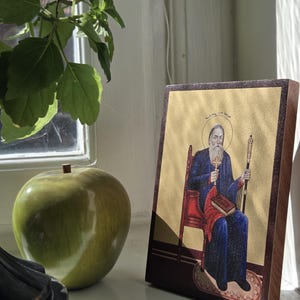 May include: A religious icon depicting a seated figure with a long white beard, wearing a blue robe and holding a staff and book. The icon is set against a gold background and framed in dark wood. A green apple and plant are in the background.