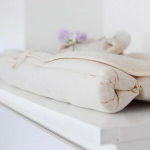 Puede incluir: Una manta plegada, de color crema, con una textura acolchada, sobre una superficie blanca. Una pequeña ramita de flores moradas descansa encima. La manta parece suave y afelpada.