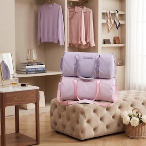 May include: Two striped duffel bags, one lavender and one pink, stacked on a tufted ottoman. The lavender bag is embroidered with "Sienna," and the pink bag with "Sophia." A wardrobe with clothing and accessories is in the background.