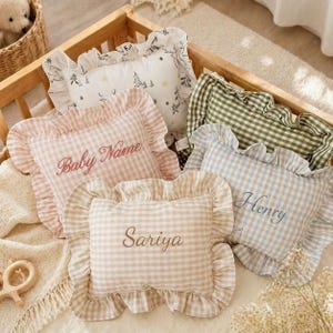 May include: Several decorative pillows with frilled edges and embroidered names. The pillows display patterns such as gingham and a winter forest design. The colour scheme includes pink, blue, green, and neutral tones, creating a soft and inviting aesthetic.