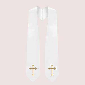 May include: White graduation stole with gold crosses embroidered near the bottom. The stole is made of a smooth, satin-like material. The background is a soft pink.