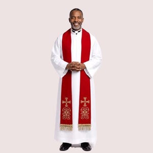 May include: A person wearing a white robe and a red stole. The stole features gold embroidered crosses and decorative patterns. The robe has long sleeves and is paired with black shoes. The person is smiling.