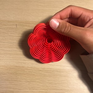 May include: A red, flower-shaped cookie cutter with a scalloped edge. The cutter has concentric rings, creating a detailed floral design. The object is held by a hand, showcasing the cutter's size and shape.