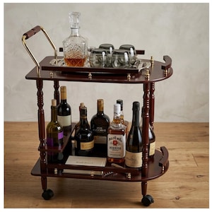 May include: A dark wood bar cart with a gold handle and accents. The top shelf holds a crystal decanter, glasses, and a silver tray. The bottom shelf displays various bottles of wine and liquor. The cart has wheels for easy movement.